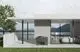 Graues Außenteil einer Wärmepumpe mit quadratischem Design, platziert auf einer Terrasse mit Blick auf Wasser und Berge, umgeben von einer modernen Architektur. Marke: Dream Maker.